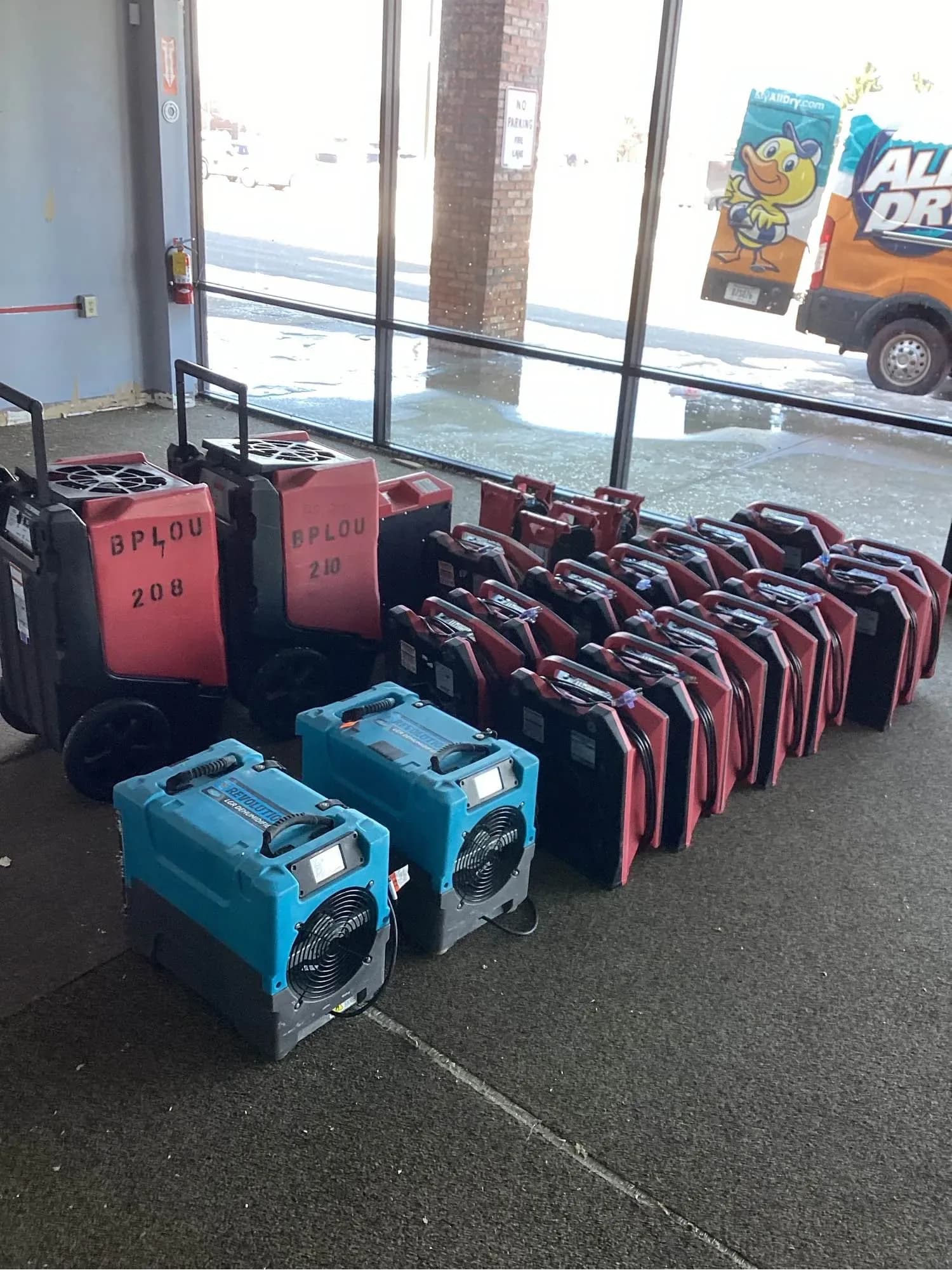 Commercial dehumidifiers and air movers staged inside a property with the All Dry van parked outside the window