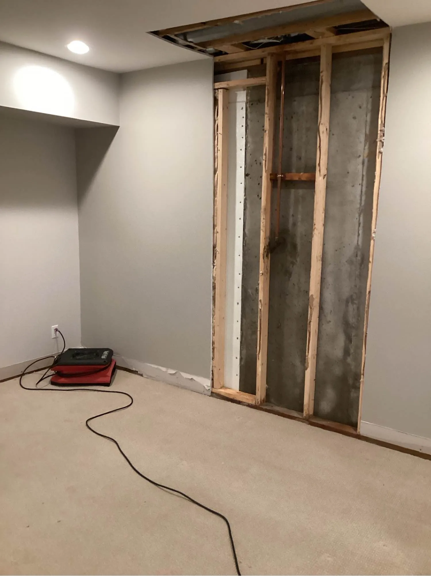 Open wall cavity with exposed studs during water damage restoration, drying fan running on the carpet nearby